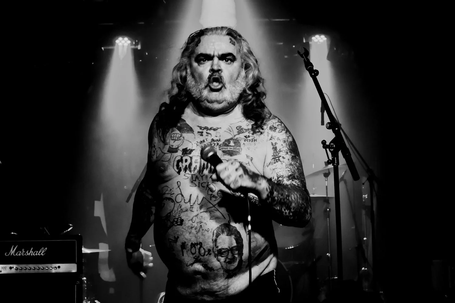 Black and white photo of a tattooed man with long hair, singing into a microphone on stage. Stage lights and musical equipment are visible in the background.