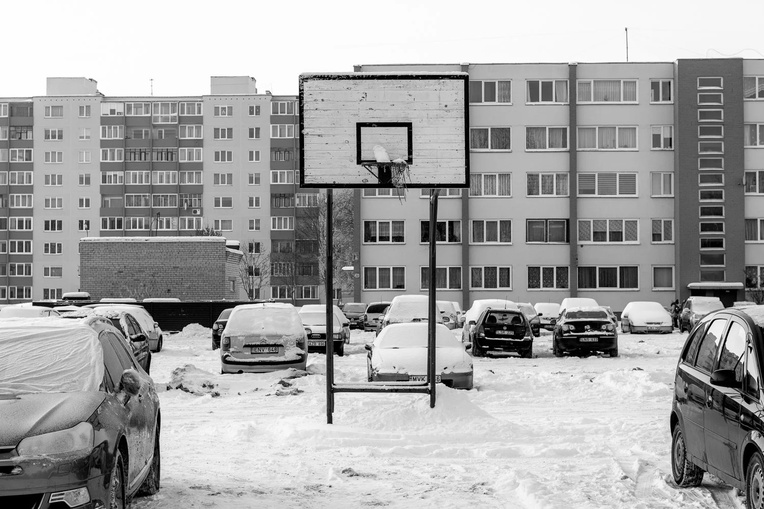 A snow-covered outdoor basketball court with a weathered backboard and hoop, surrounded by parked cars and multi-story residential buildings in the background, in winter.