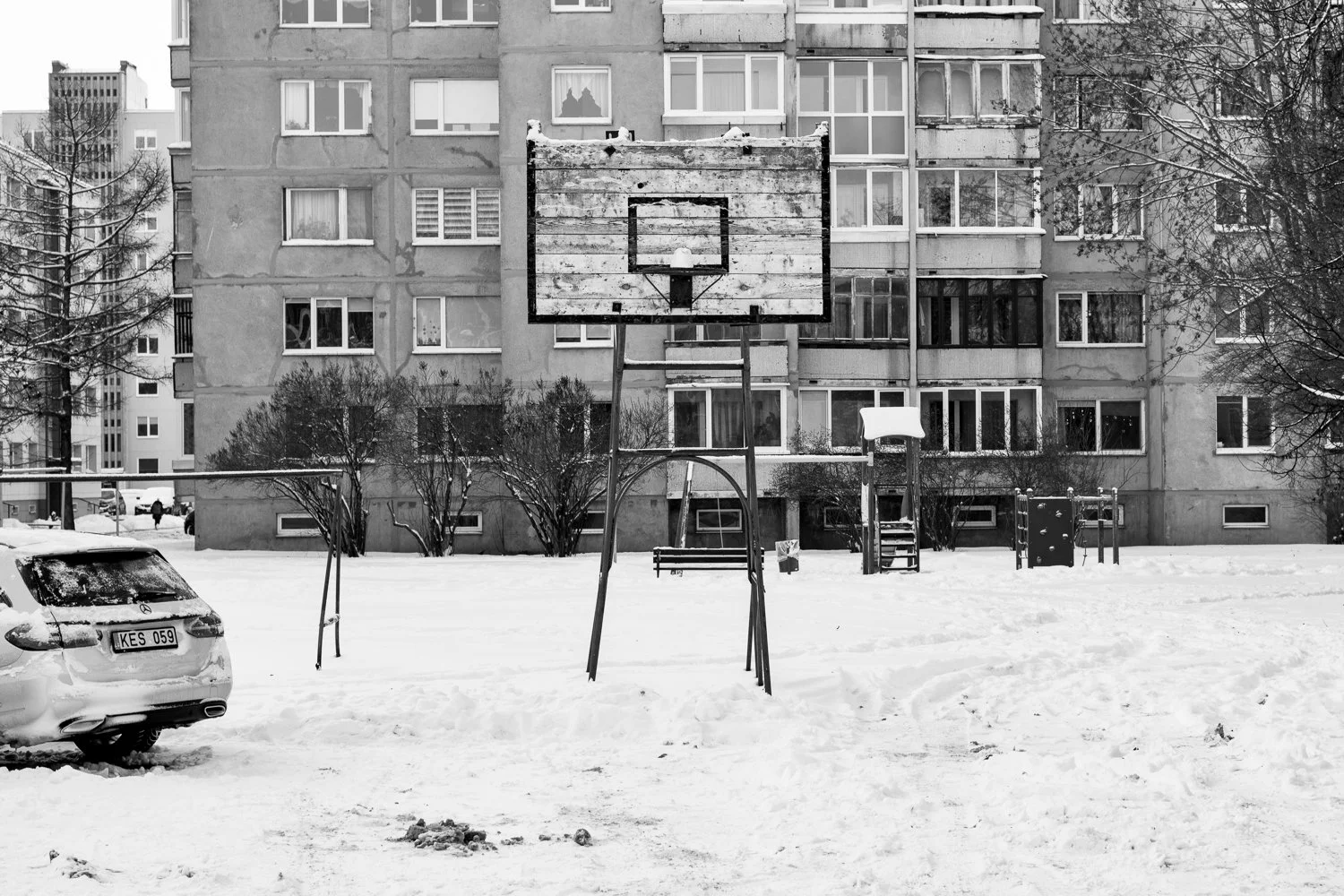 Snow-covered basketball court in front of an apartment building with leafless trees and a parked car.