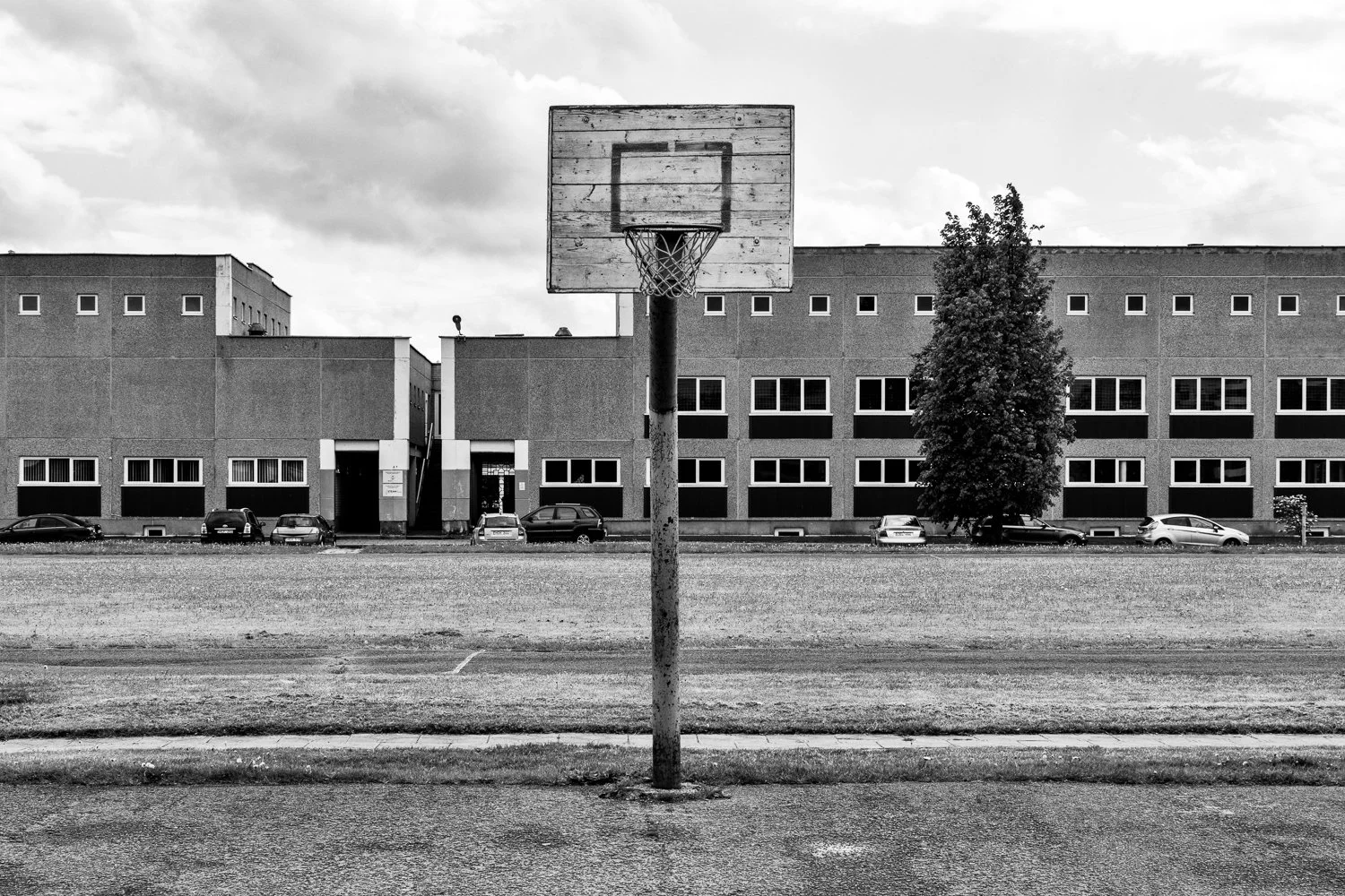 Black and white photo of an outdoor basketball hoop standing on a paved area in front of a large multi-story building with parked cars and a large tree.