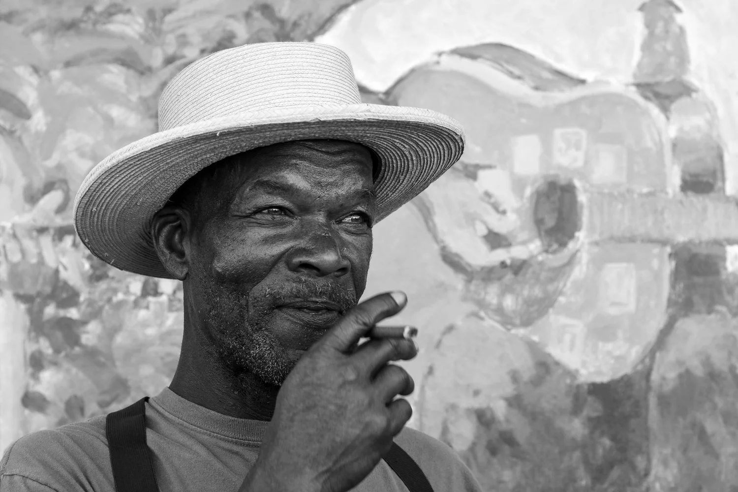An older man with dark skin wearing a wide-brimmed hat and a t-shirt, holding a cigarette near his mouth, with a large abstract painting or mural in the background.