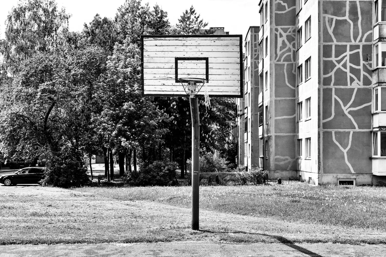 An empty outdoor basketball hoop on a court in front of a residential apartment building with trees in the background in black and white.