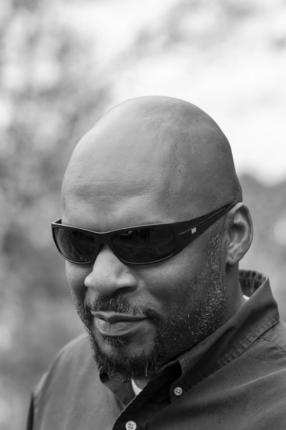 A black-and-white close-up portrait of a bald man wearing dark sunglasses and a collared shirt.