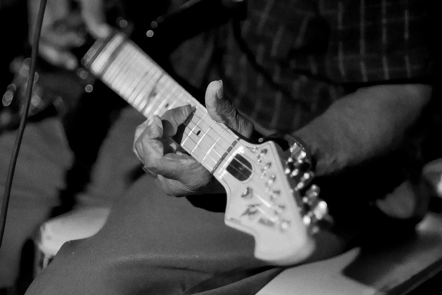 Close-up of a person's hand playing an electric guitar, wearing a plaid shirt, in black and white.