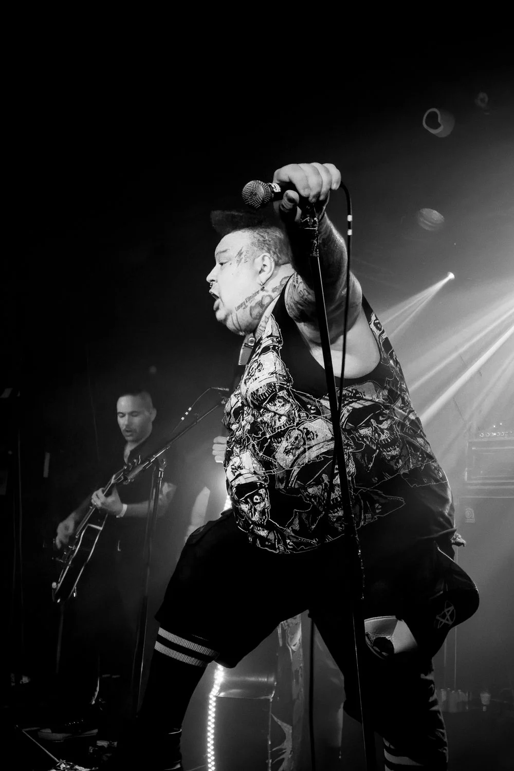 A black and white photo of a performer holding a microphone on stage, with another musician playing guitar in the background, under stage lights.