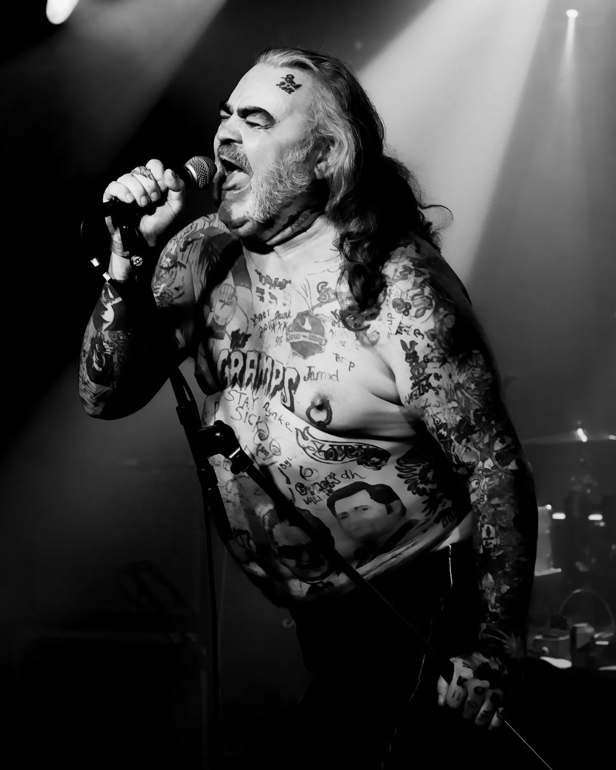 A tattooed male singer with long hair singing into a microphone on stage, with dramatic lighting.
