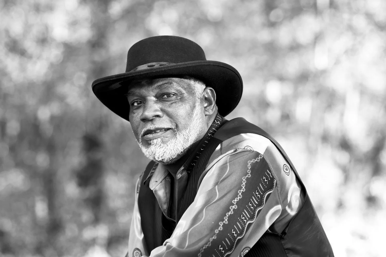 A black and white portrait of an older man with a white beard wearing a wide-brimmed hat and patterned shirt, outdoors with a blurred background.