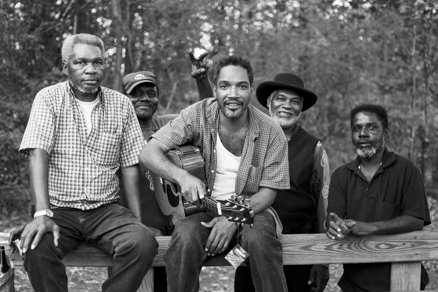 Black-and-white photo of six men outdoors, one man in the center holding a guitar. The men are sitting and standing around a wooden bench, with trees in the background.