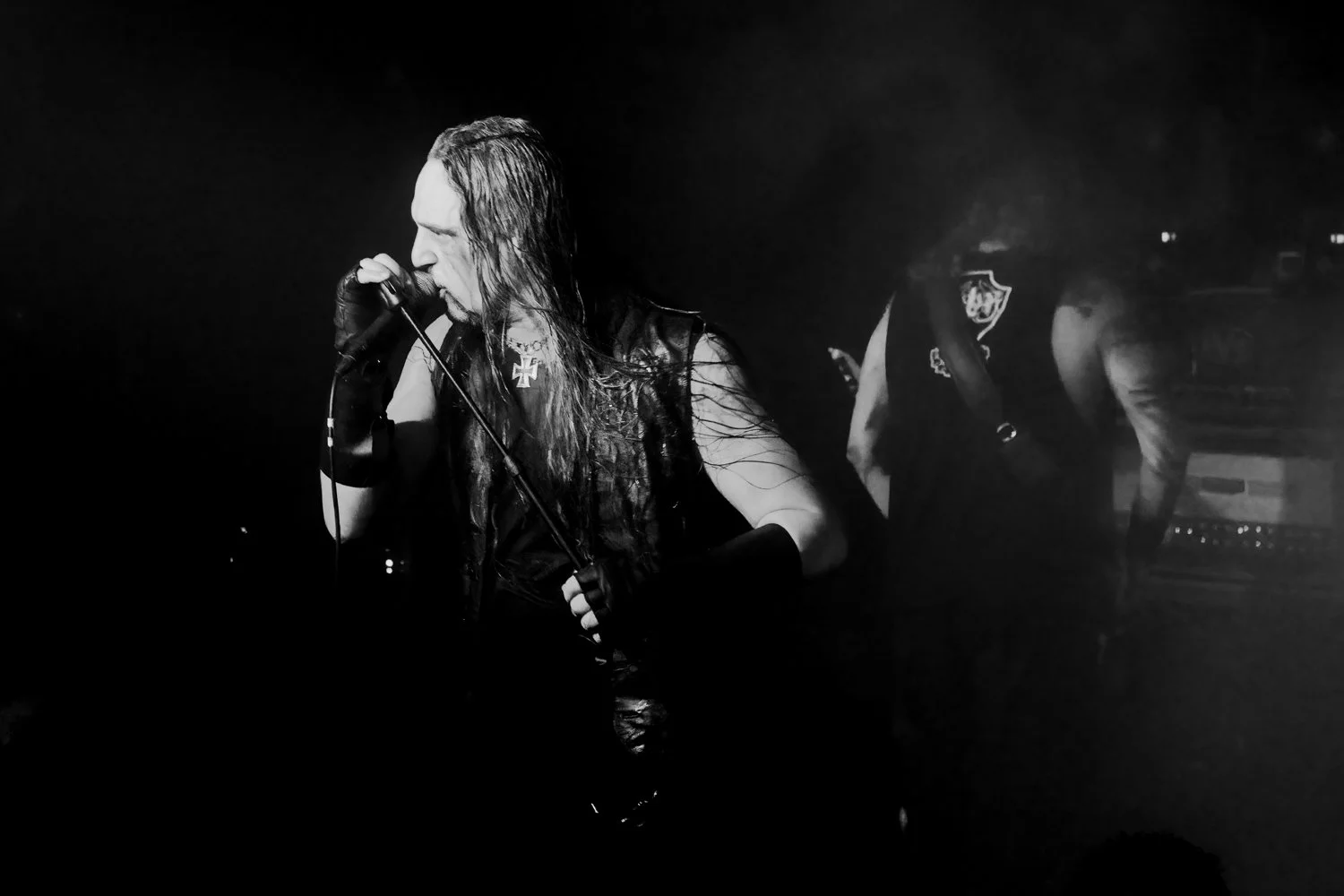 Black and white photo of a male singer with long hair, wearing a sleeveless shirt with a cross patch, singing into a microphone on stage, with another band member in the background.