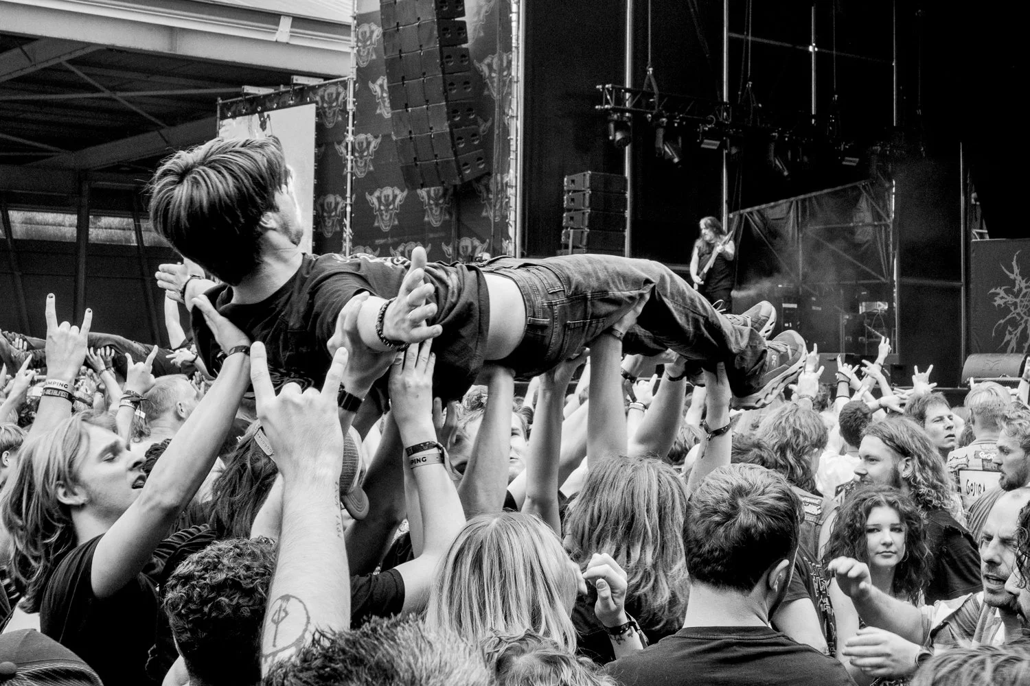 Black and white photo of a crowd at a concert. A person is crowd surfing, lying horizontally on people's hands, with their arms outstretched. The stage is visible in the background, with a guitarist playing.