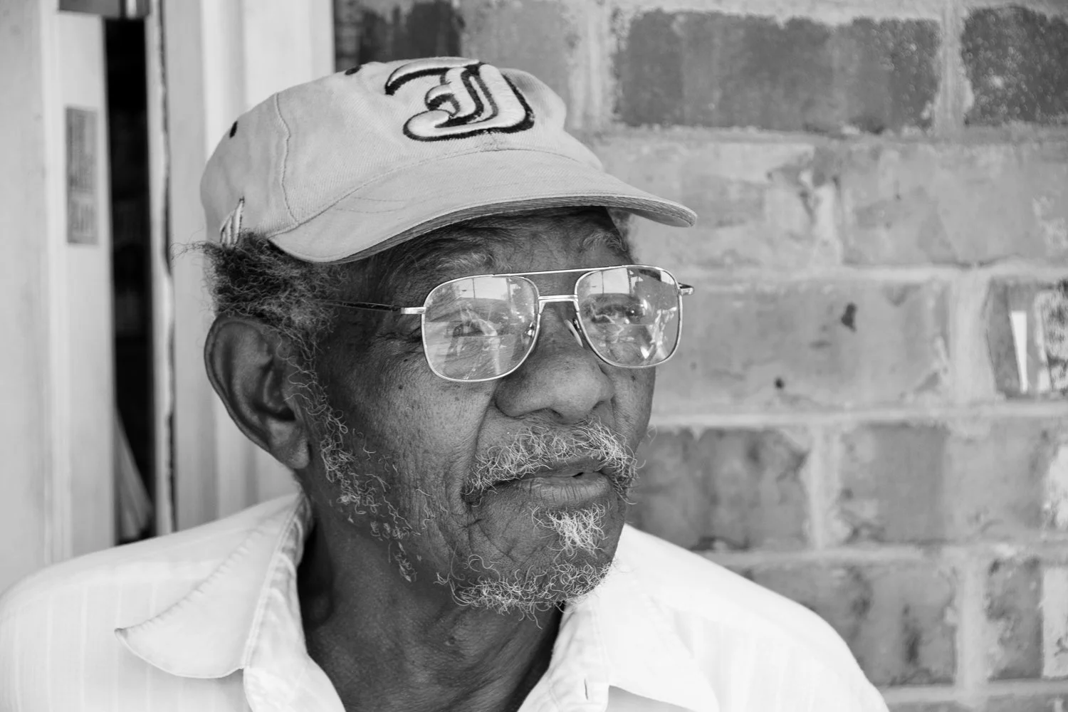 Black and white photo of an elderly man wearing glasses, a baseball cap, and a white shirt, sitting against a brick wall.