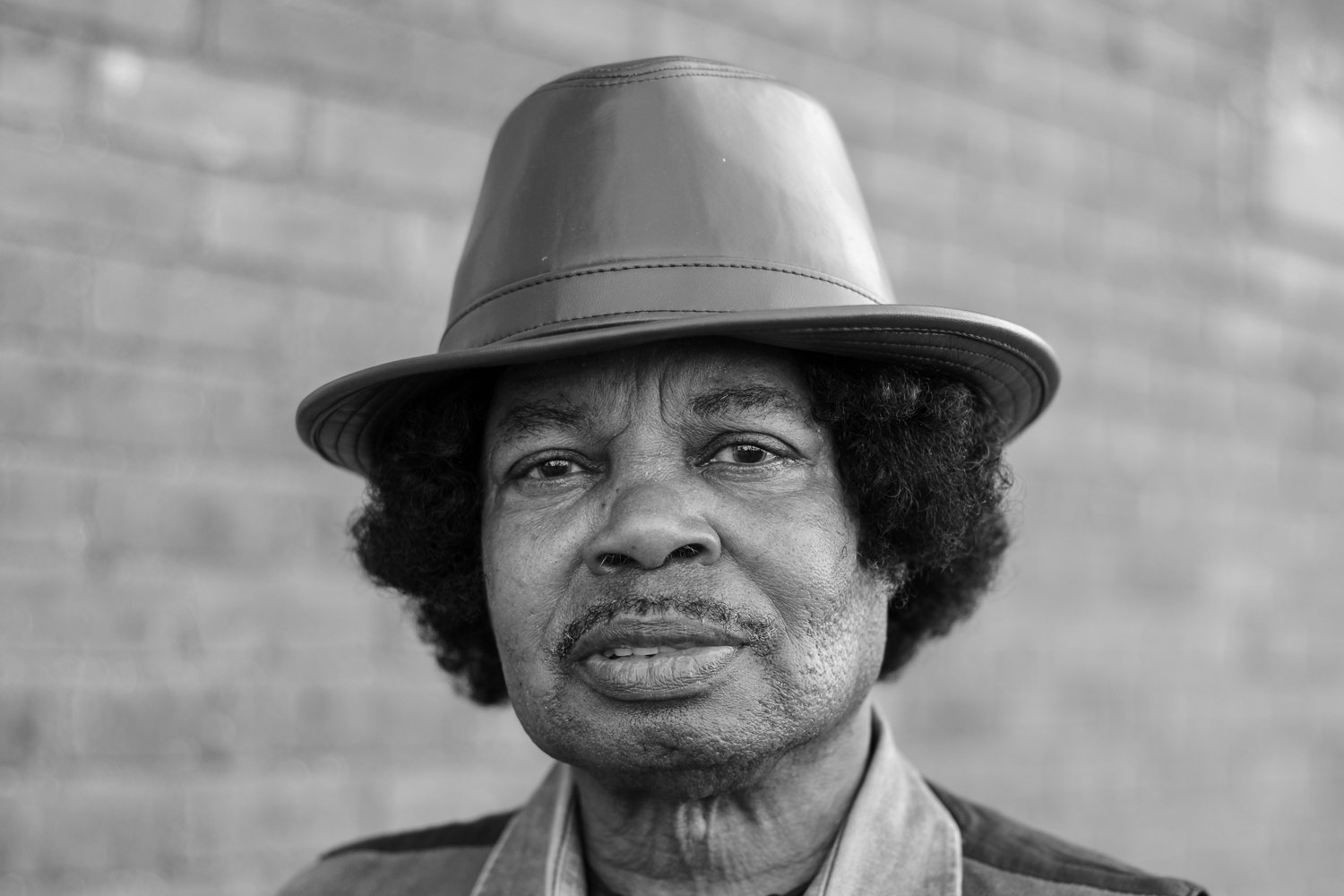 A black and white portrait of a man wearing a leather fedora hat, with a brick wall in the background.