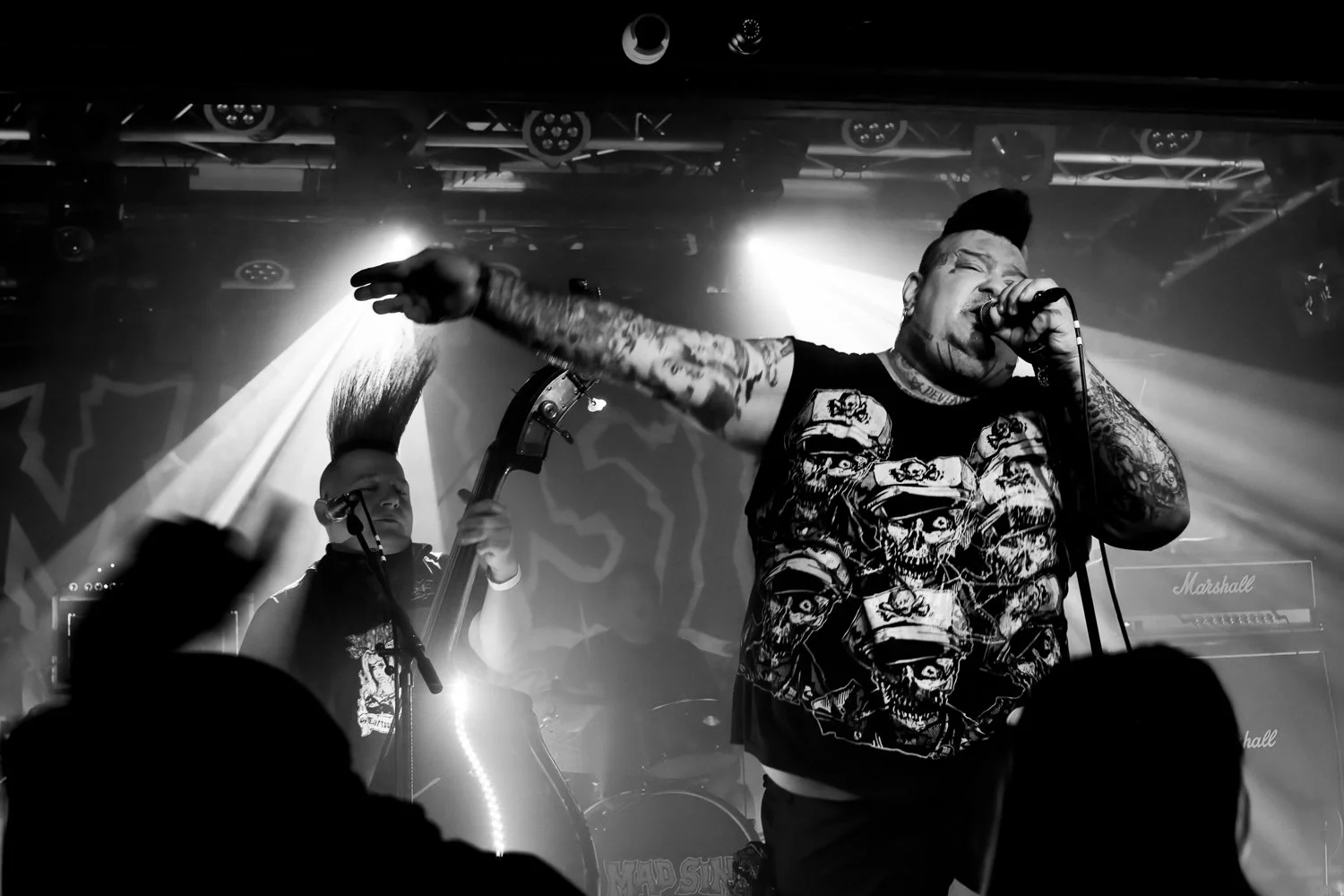 Black and white photo of a live music performance featuring a vocalist with tattoos, a sleeveless shirt with skull designs, holding a microphone with one hand while the other arm is extended, and a bassist with a mohawk hairstyle playing the bass gui