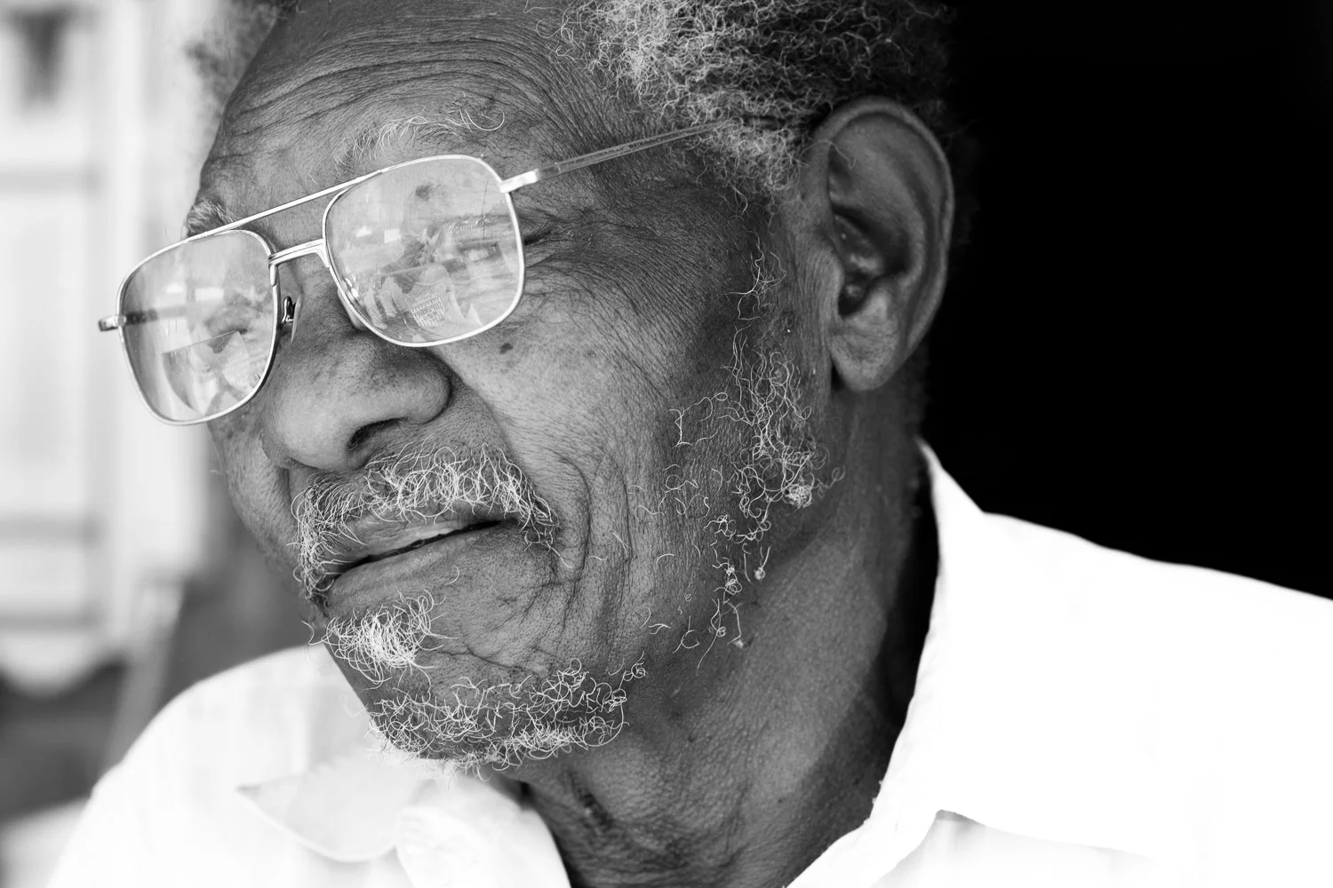 Close-up black and white photograph of an elderly man with glasses and a beard, wearing a white shirt, looking to the side.