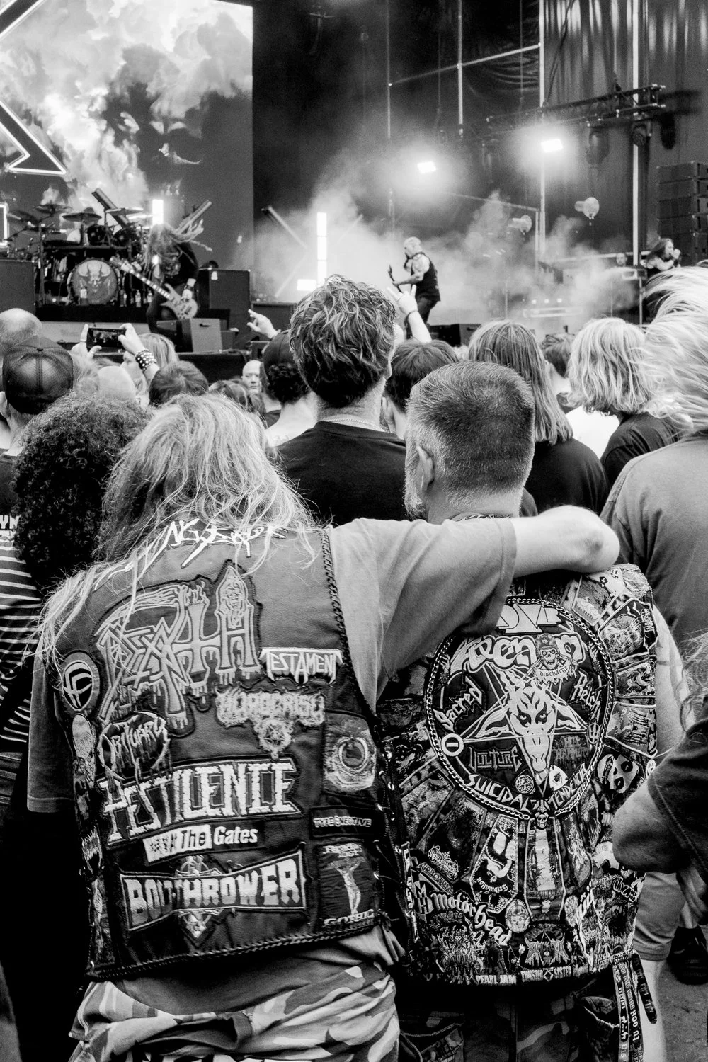 Concert scene with audience watching a band perform on stage, with some people capturing the moment on their phones. The audience members are dressed in punk and metal band patches and attire.