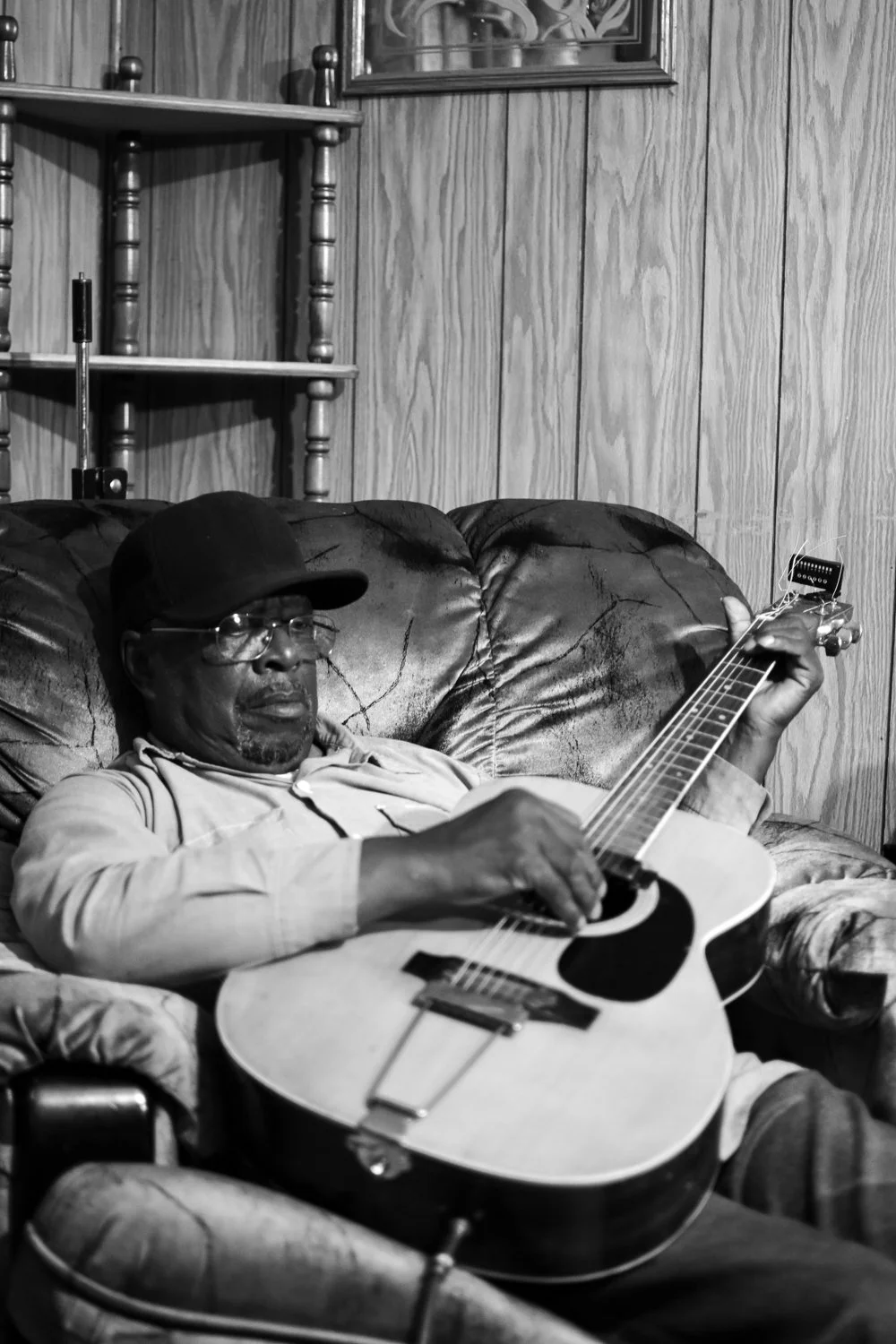 A man is sitting on a worn leather couch, playing an acoustic guitar, wearing glasses, a cap, and a light-colored shirt, in a room with wooden paneling on the wall.