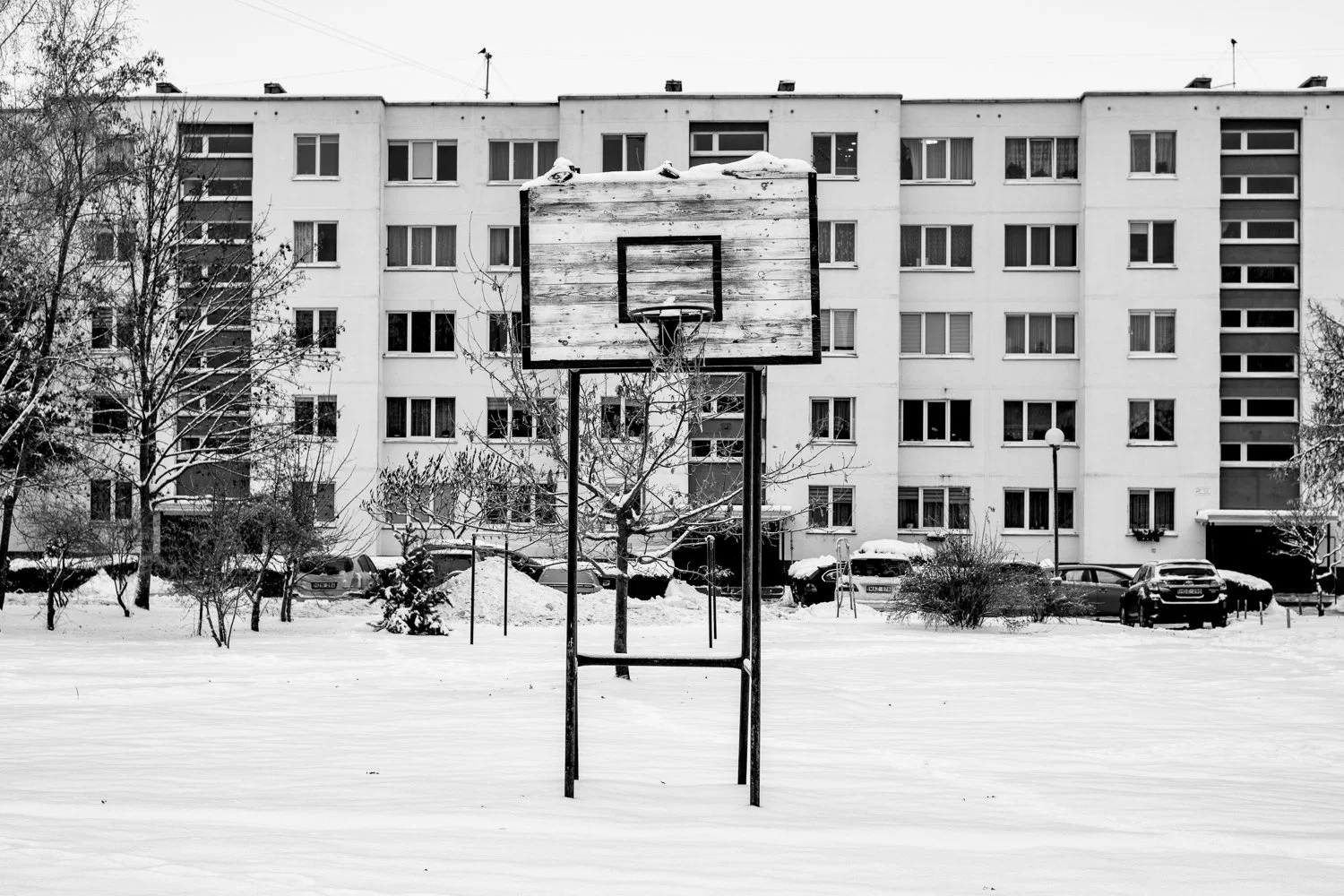 A deserted outdoor basketball court covered in snow, with a weathered basketball hoop in the foreground and a residential apartment building in the background.