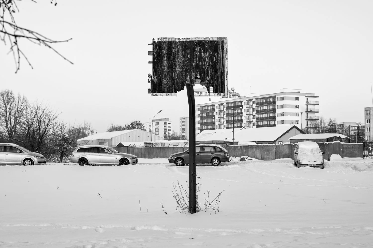 A black-and-white photo of an empty outdoor basketball hoop on a snowy field, with a row of parked cars and modern apartment buildings in the background.