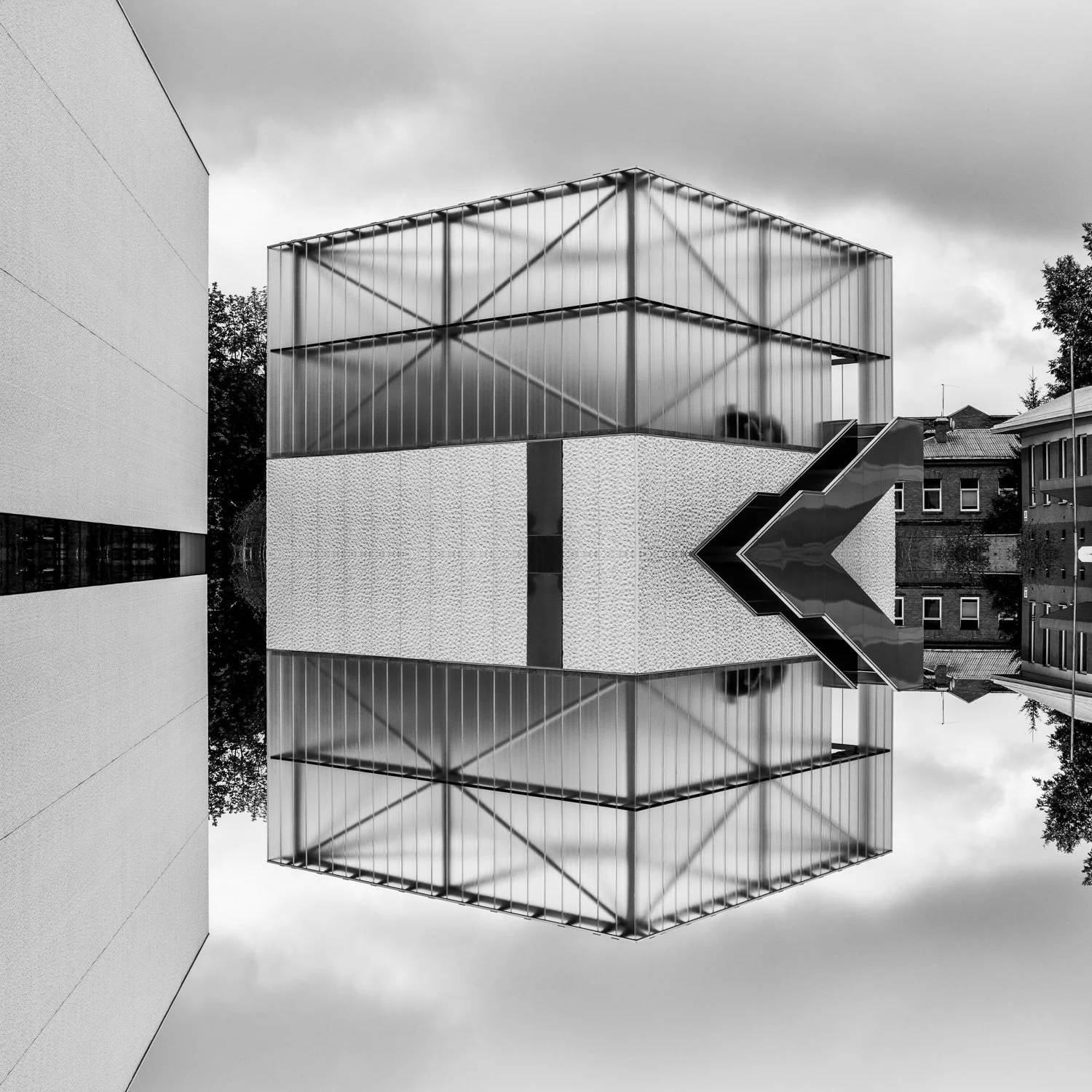 A modern building with geometric design, featuring a large, illuminated glass box on top and a reflective surface below, creating a mirror image of the structure and surroundings.