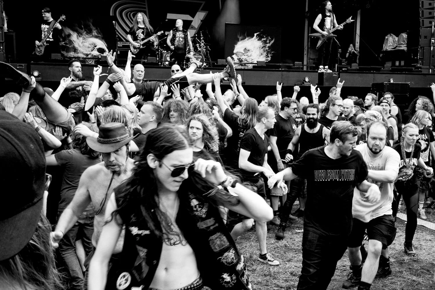 A black and white photo of a lively music concert, with people crowding the area in front of the stage. Some audience members are dancing and lifting each other, while the band performs on stage in the background. The band members are playing guitars