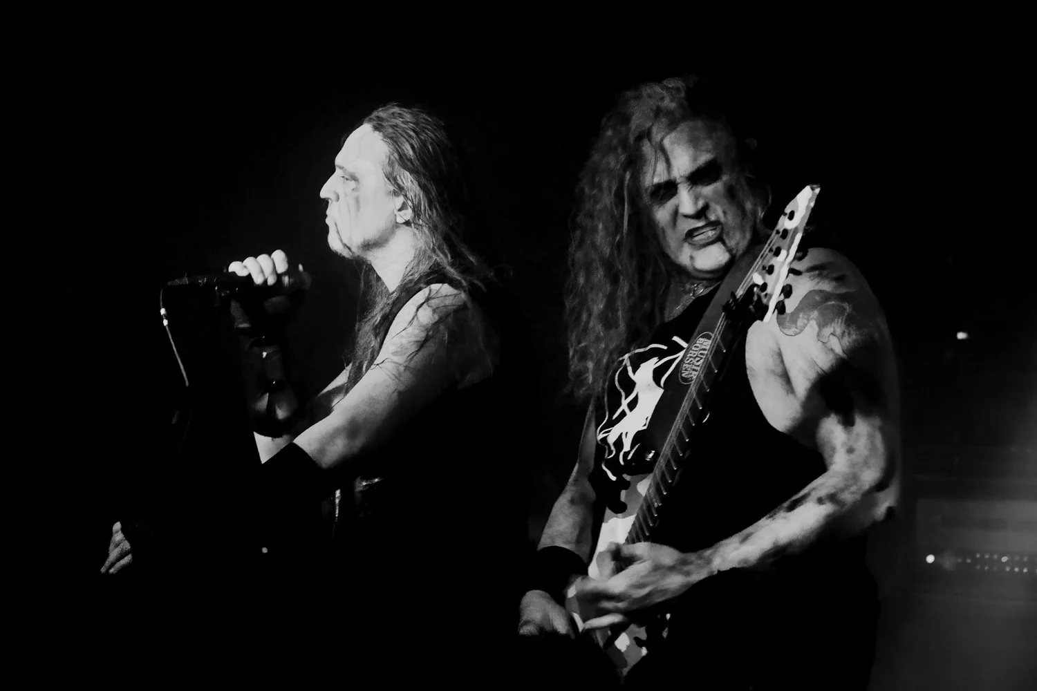 Black and white photo of two male rock musicians performing passionately on stage. The singer is on the left, with long hair, singing into a microphone. The guitarist is on the right, with long hair and tattoos, playing an electric guitar and making 