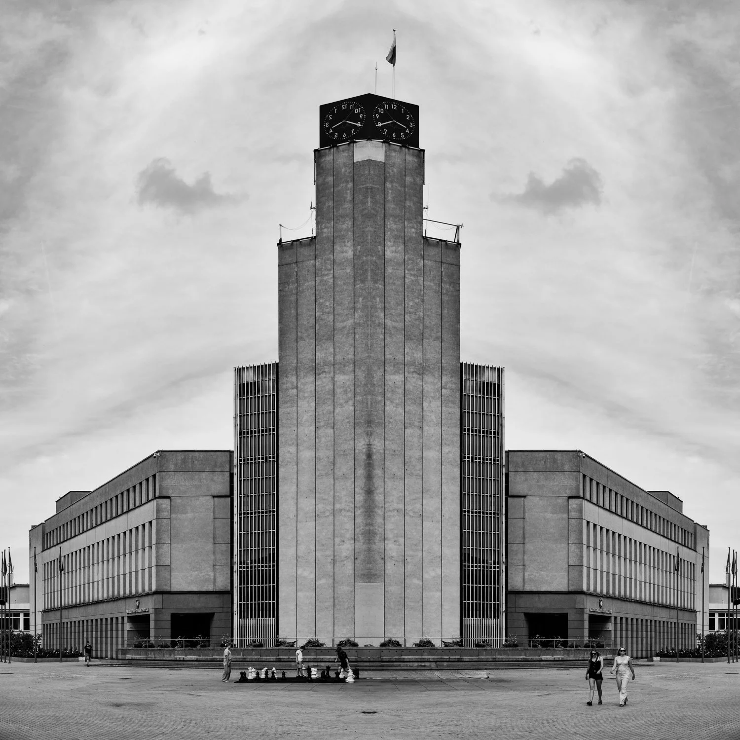 A large government building with a central tower featuring a clock on top, surrounded by cloudy sky, with people walking in front.