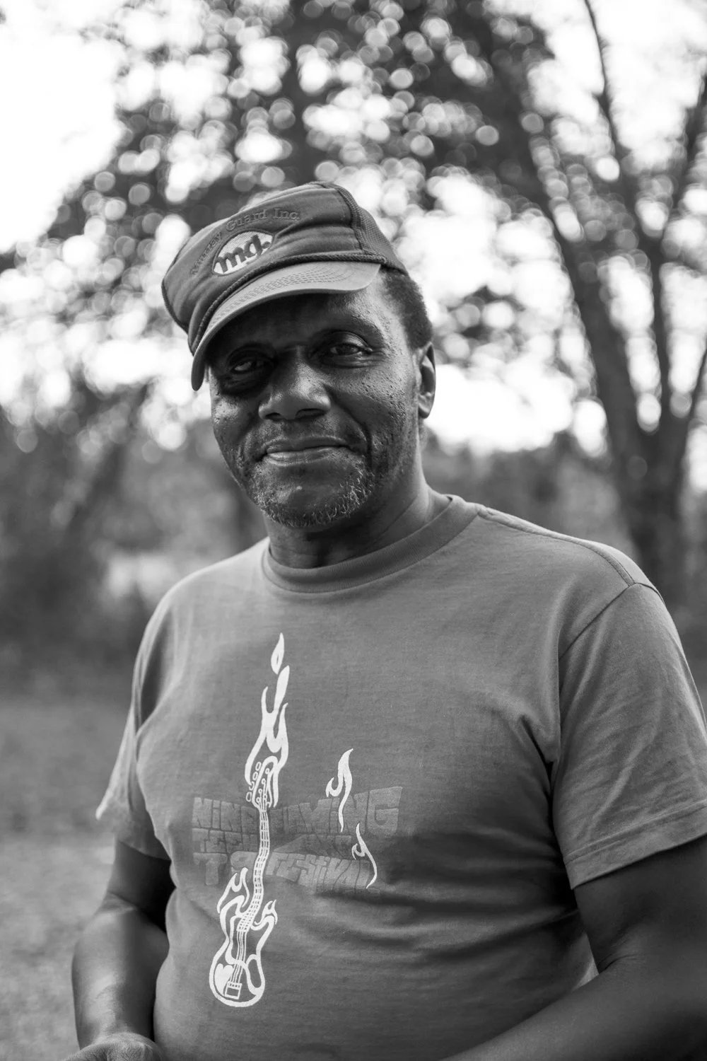 A middle-aged man standing outdoors in a park or forest, wearing a cap and a t-shirt with a guitar and flames graphic, smiling softly with trees blurred in the background.