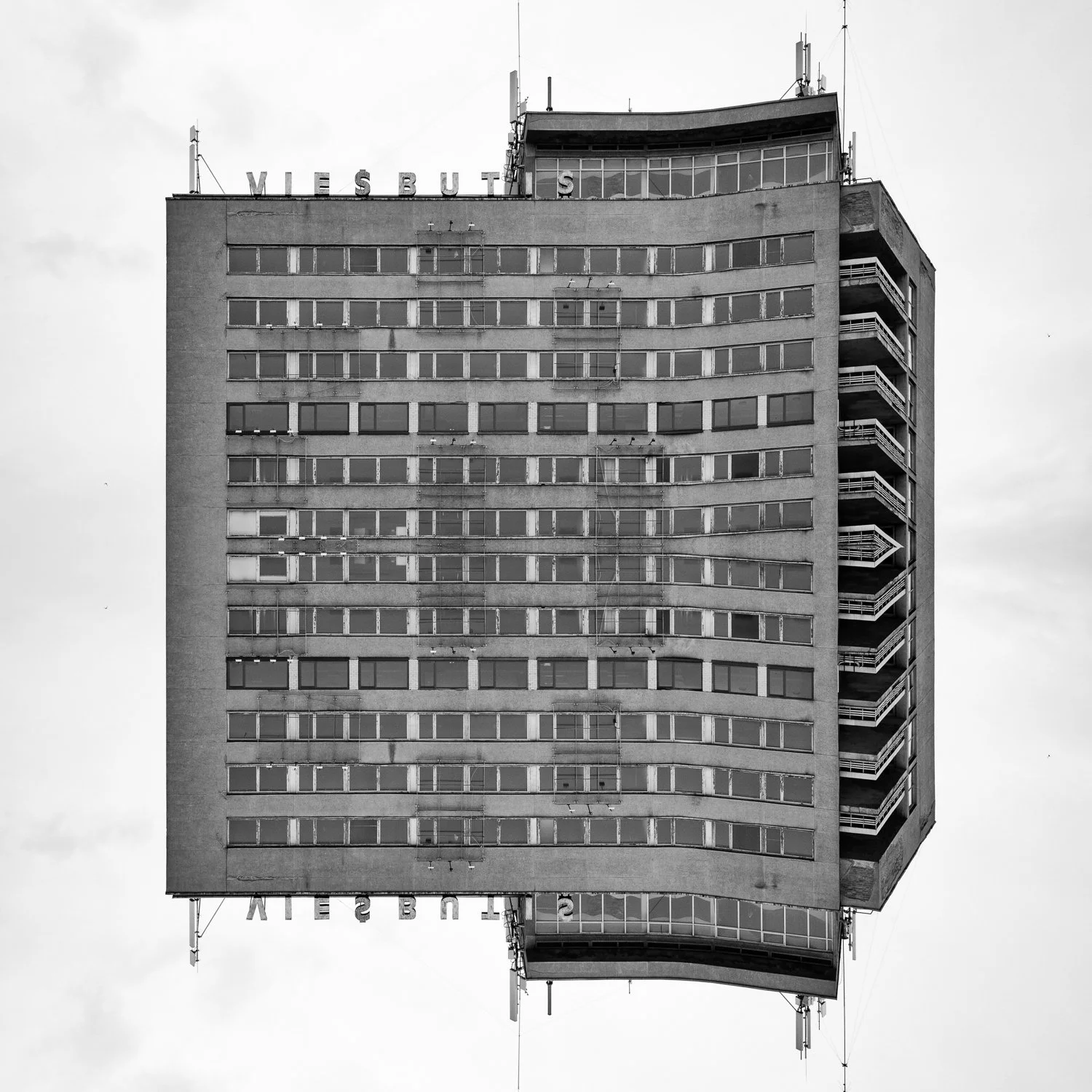 A black and white photo of a tall, multi-story building with many windows and balconies, with the word 'VIESBUTIS' partially visible at the top.
