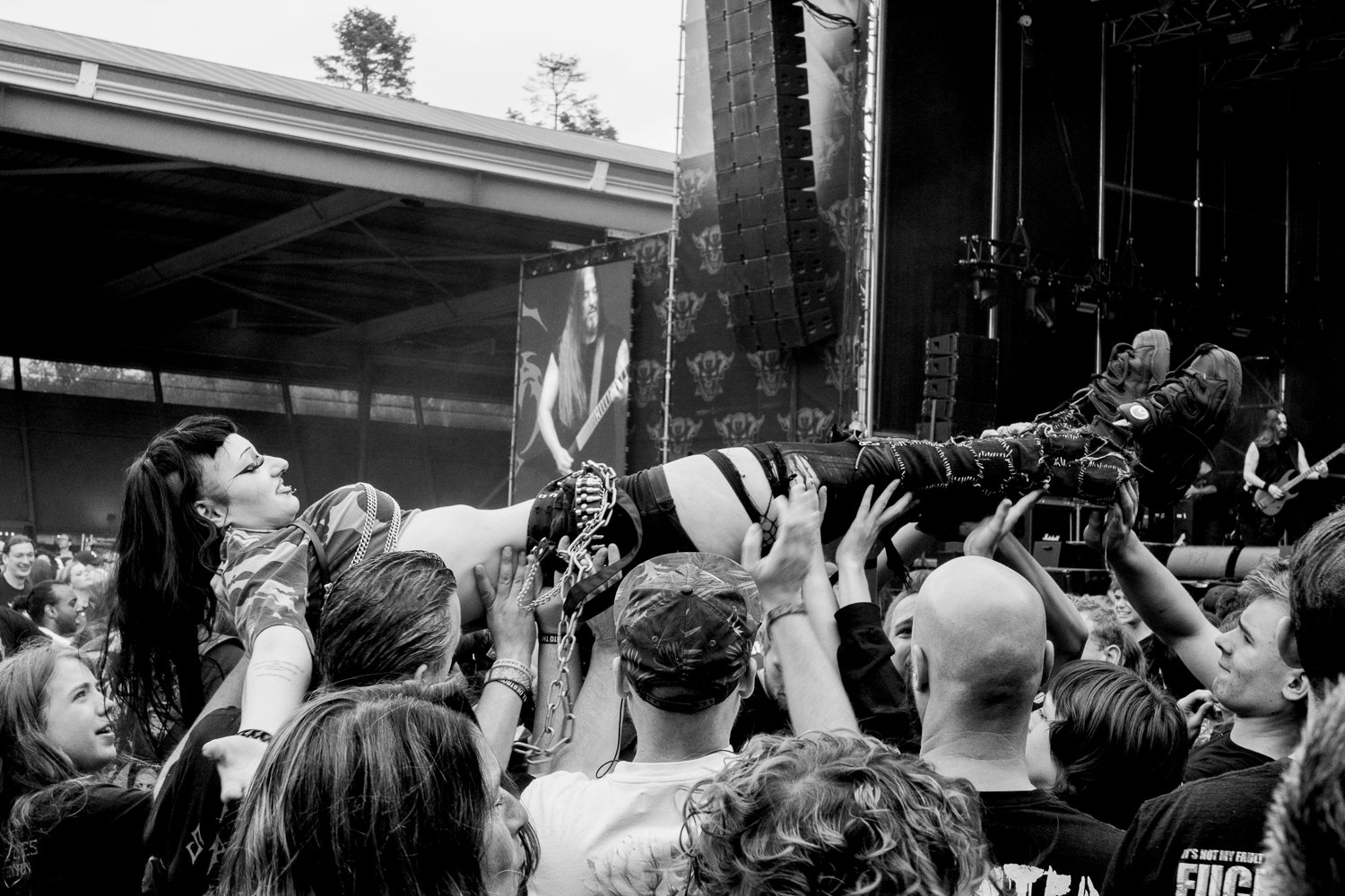 A crowd at a concert lifting a woman onto their hands near the stage, with musicians performing in the background.