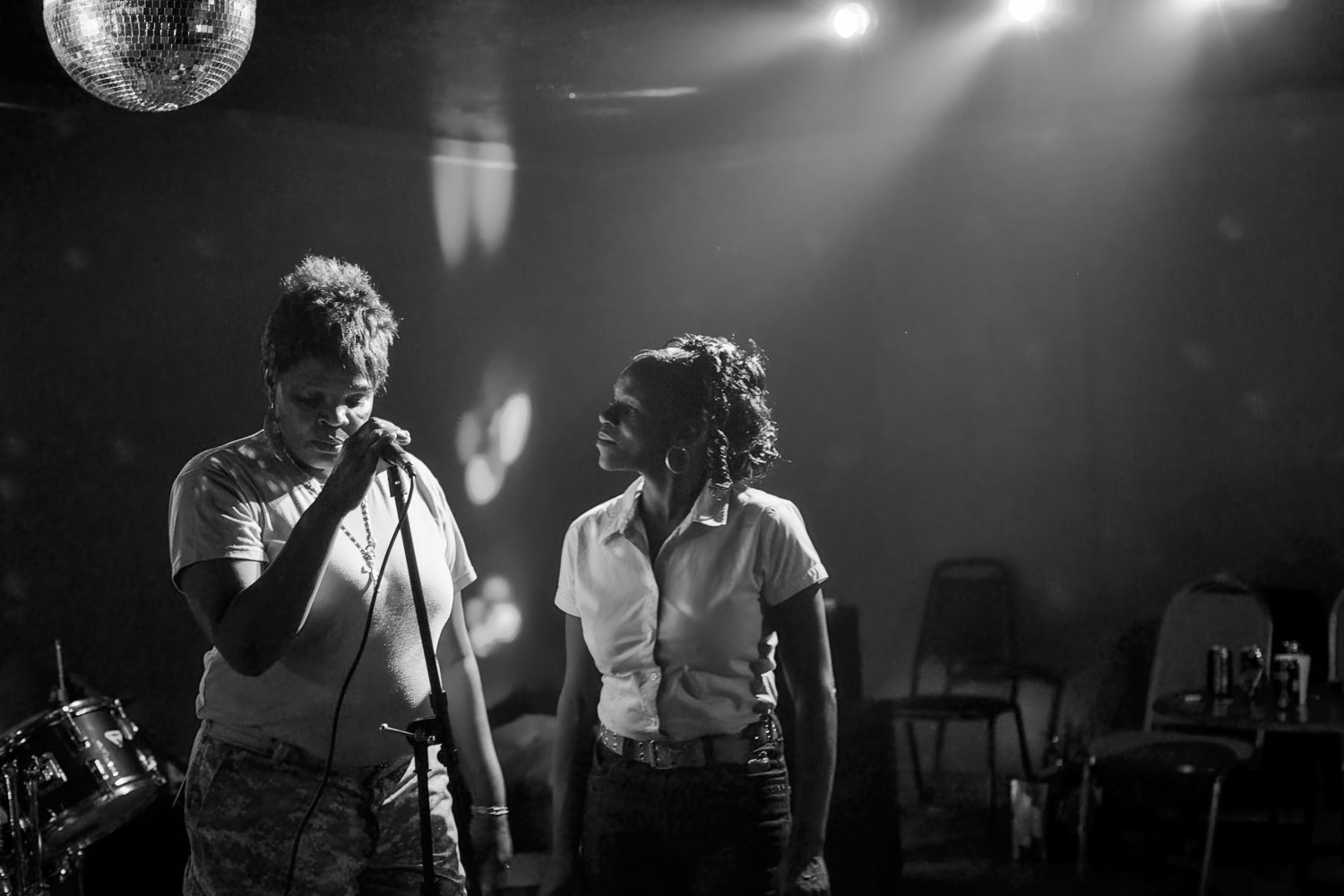 Two women standing in a dimly lit room with a disco ball hanging from the ceiling, one holding a microphone and the other looking at her, with chairs and a drum set in the background.