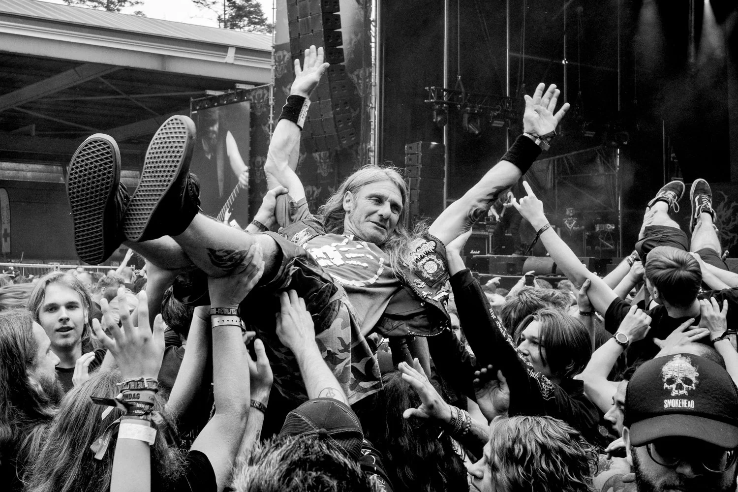 A man crowd surfing at a concert, surrounded by fans, in an indoor venue with a stage and lighting in the background.