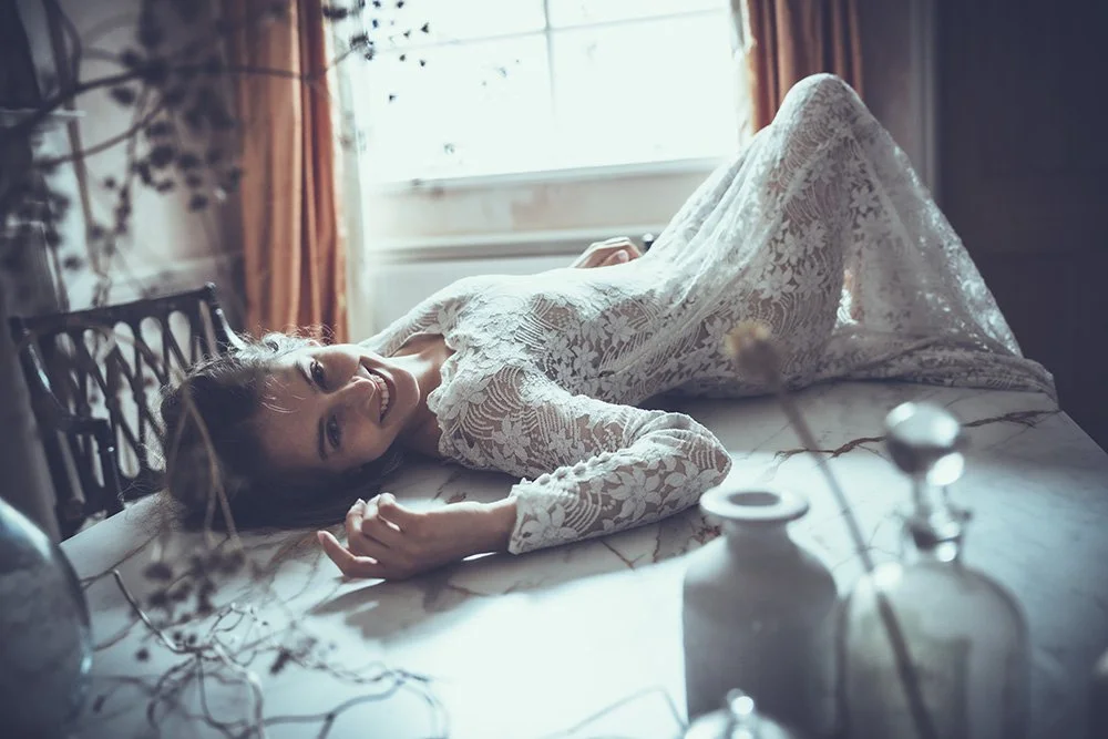 Bride lying on a wooden table in a lace bridal gown, smiling, in a room with natural light and decorative bottles.