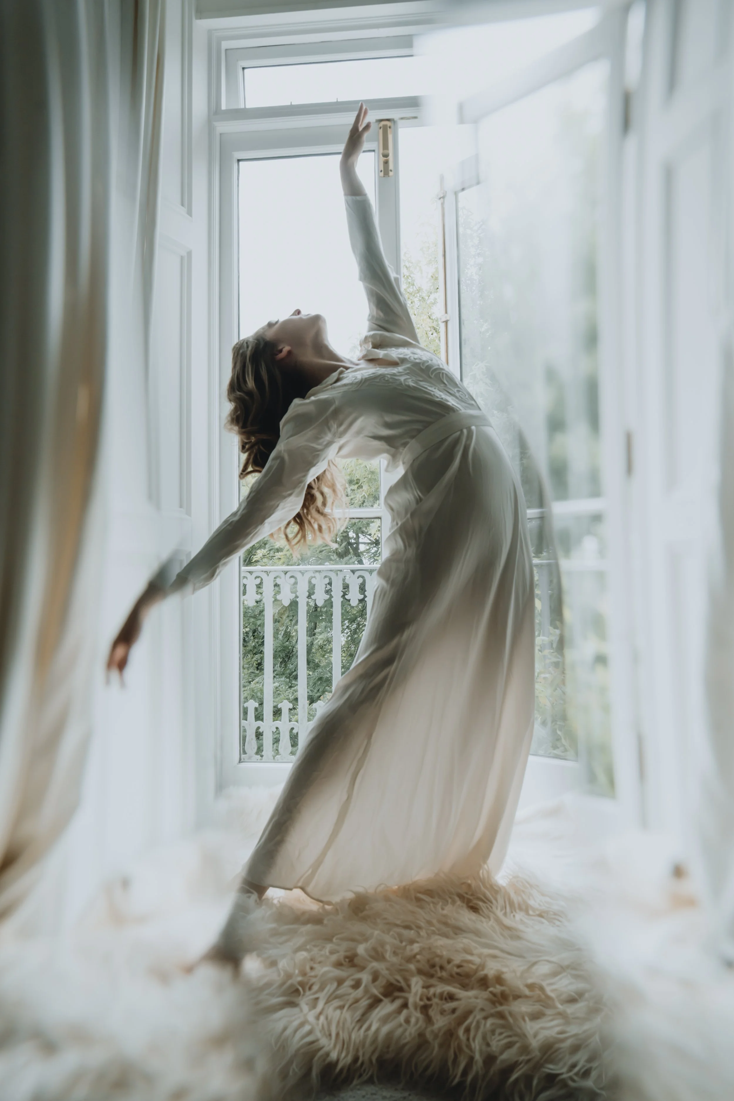 A woman in a white wedding dress by a large window with a balcony, with sunlight coming through the window.