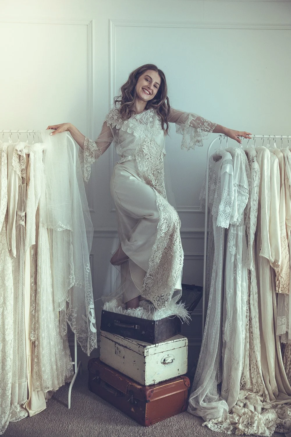 A bride dressed in vintage lace and satin Tuesday's Bride wedding gown standing on a stack of three antique suitcases, surrounded by racks of white and cream lace wedding dresses. She is smiling.