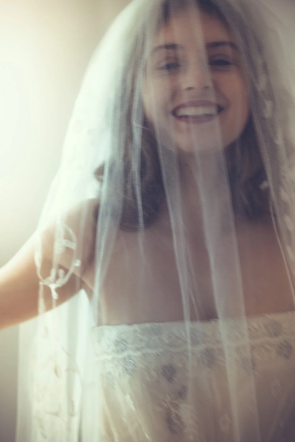 A woman smiling behind a vintage bridal veil, likely on her wedding day.