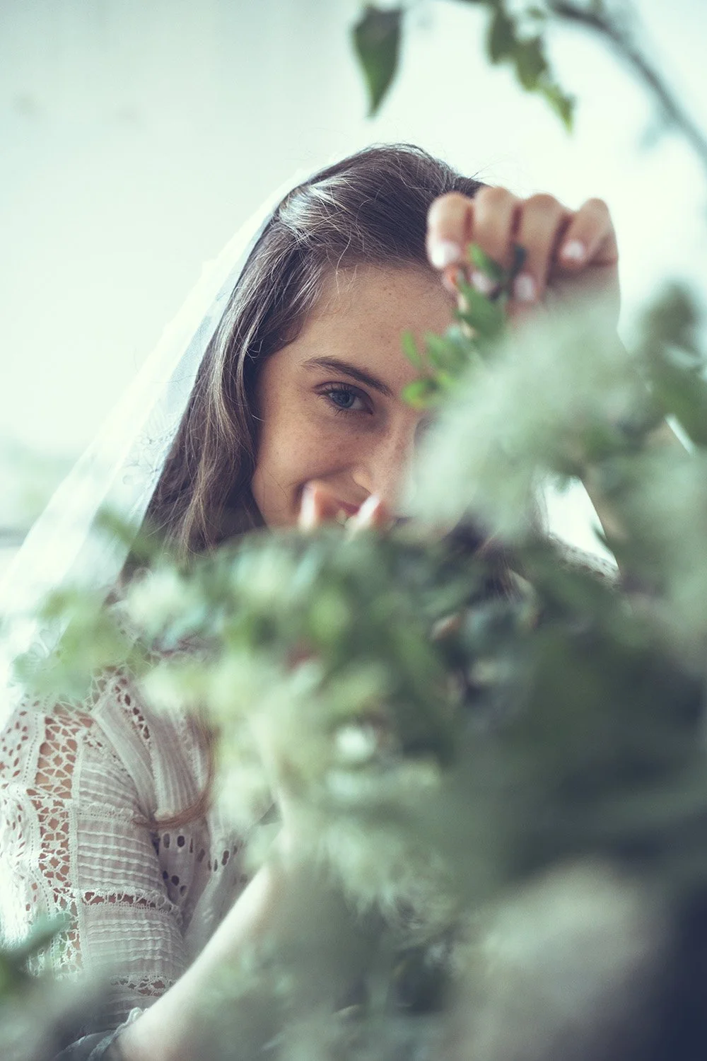 A young bride with long brown hair and blue eyes smiling, partially hidden by greenery, wearing a white lace wedding dress and a white veil.
