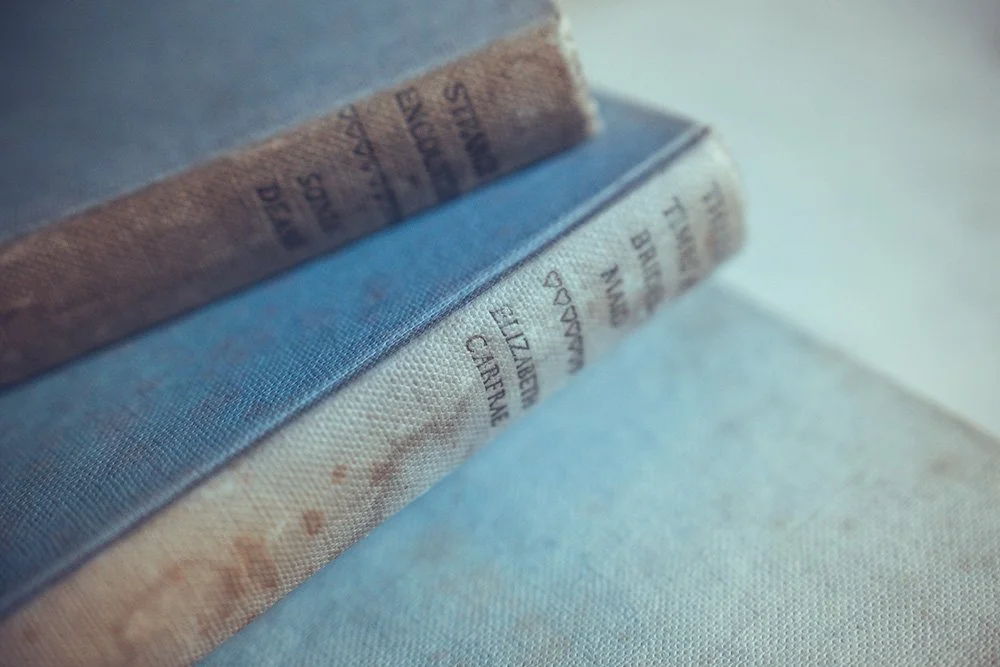 Close-up of two stacked books with visible spines, one with a light brown spine and the other with a white spine, resting on a light-colored fabric surface.
