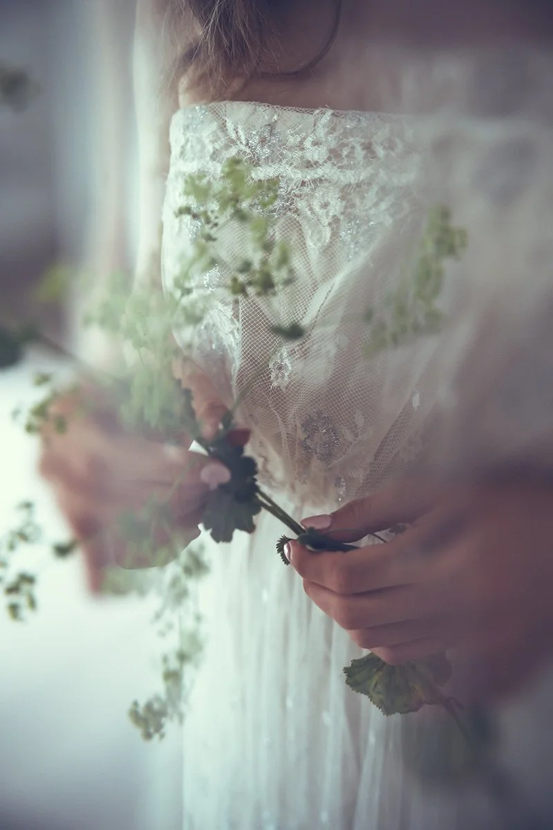 Bride holding a small bouquet of flowers, partially covered by lace fabric in a soft, muted light.