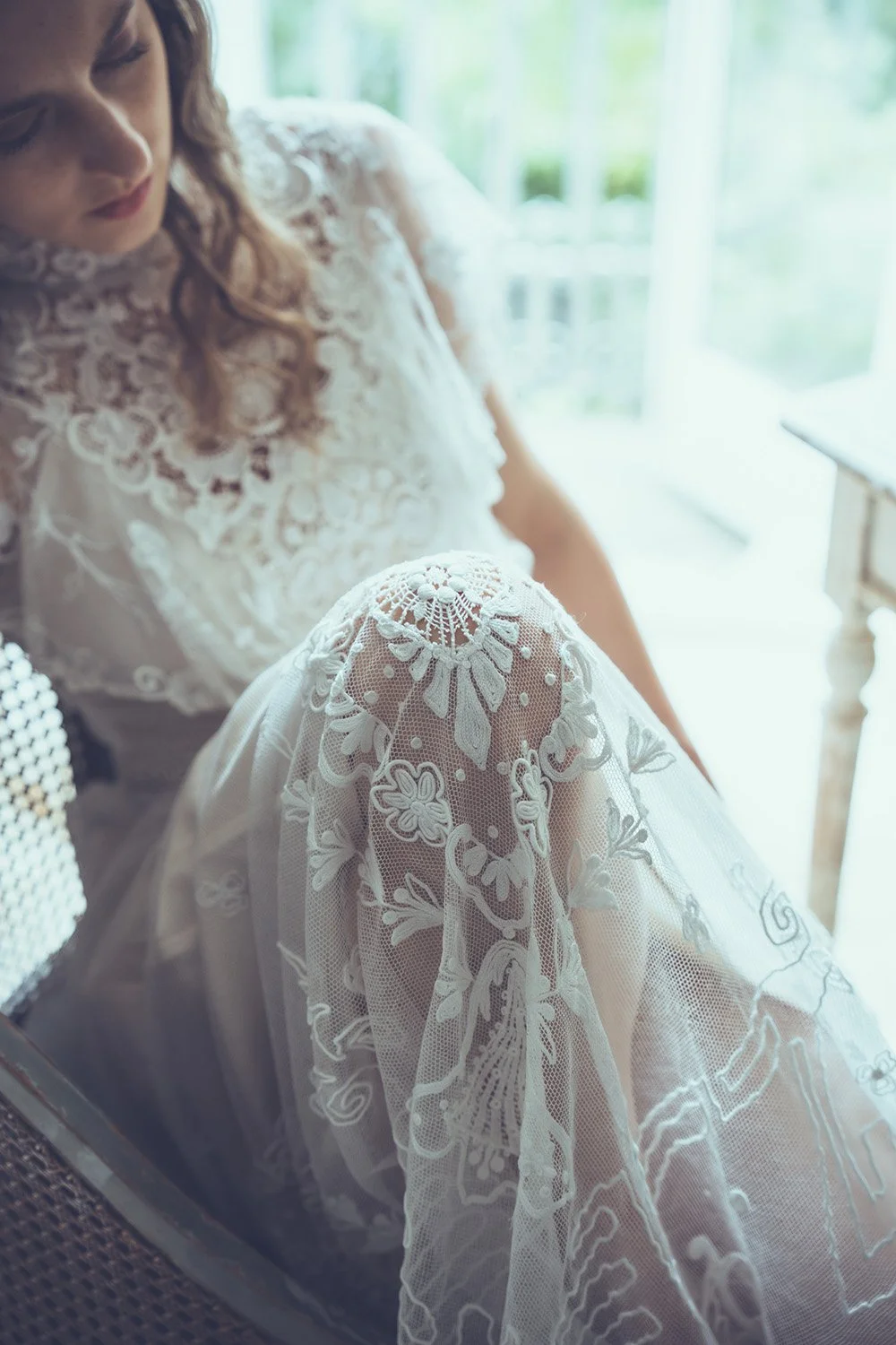 A bride wearing a white lace wedding dress sitting near a window, with sunlight illuminating her from behind, and a detailed lace pattern on her wedding gown's shoulder and sleeve.