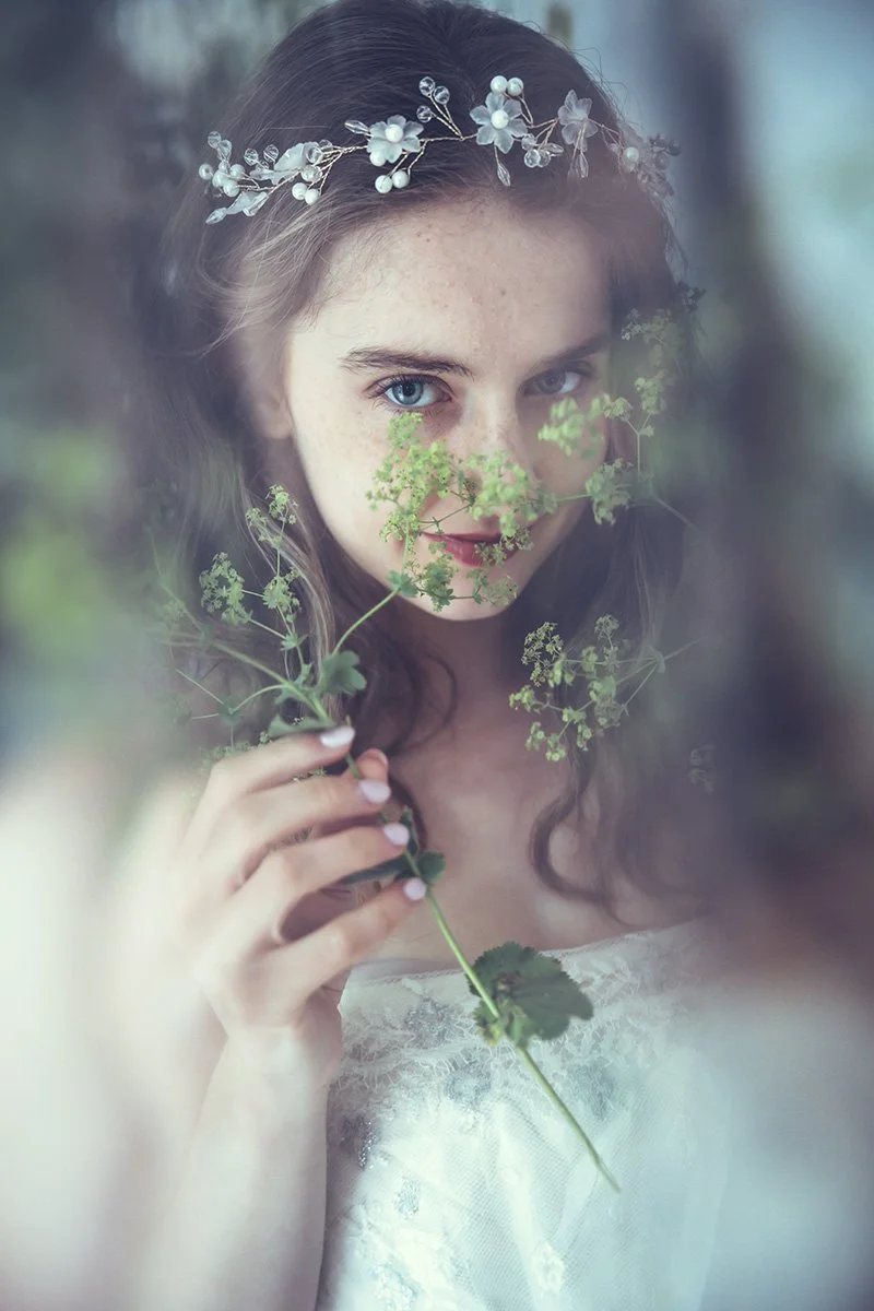 A bride-to-be with blue eyes and long dark hair wearing a  Tuesday's Bride wedding floral headband, holding a sprig of green plants, with a soft, blurred background.