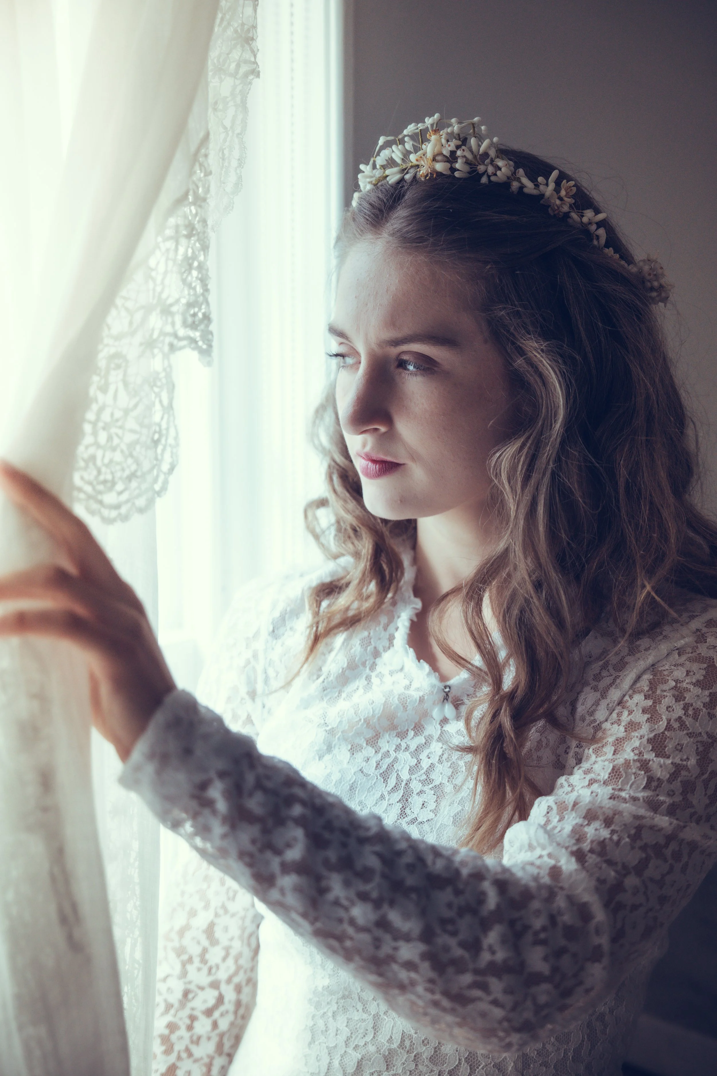 A bride with long, wavy hair wearing a floral wedding headdress and a lace bridal gown looks out the window while holding a curtain.