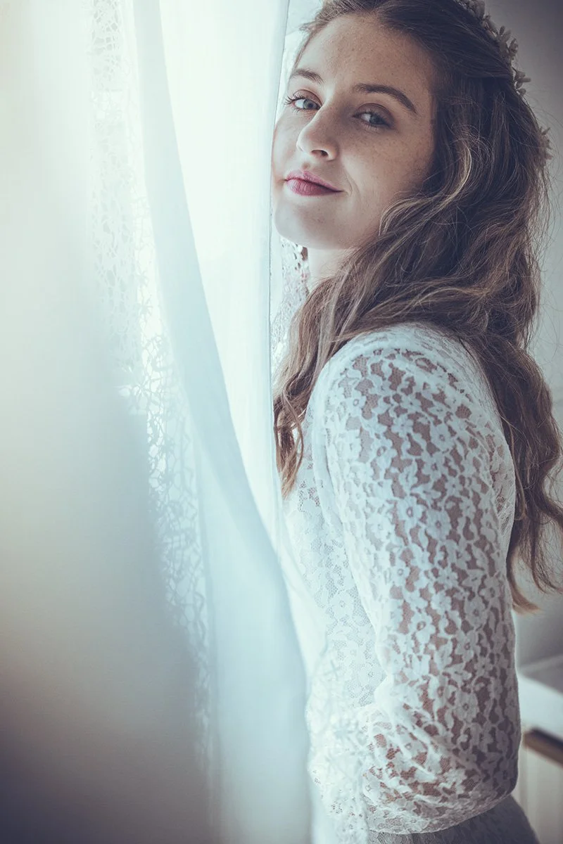 Young bride with brown hair and freckles looking at camera through a sheer white curtain.