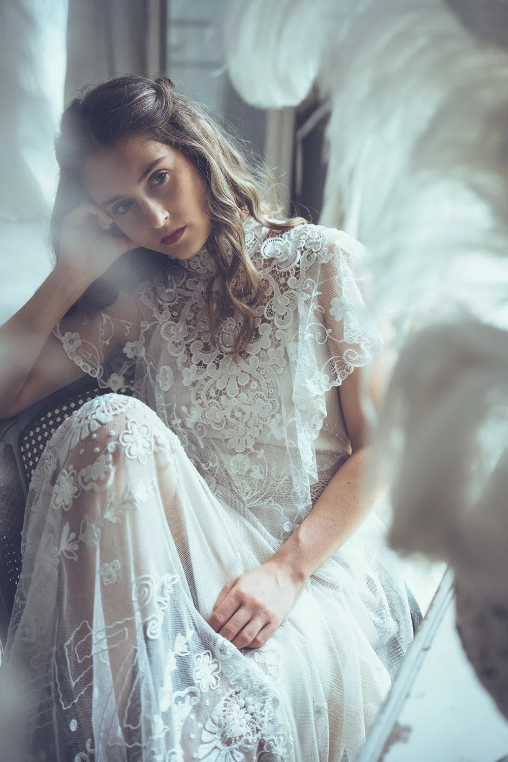 Bride with wavy hair wearing a lace wedding dress sitting near a window with feathers in the foreground