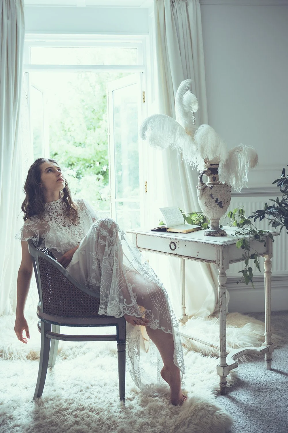 A  with long dark hair in a sheer white lace Tuesday's Bride wedding dress sits barefoot in a vintage chair near a large open window with white curtains. She looks upward. The room is bright with natural light and decorated in soft, neutral tones.