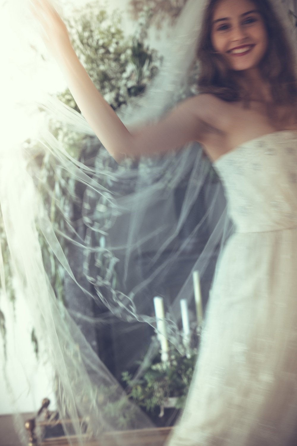A woman with long brown hair wearing a white one-of-a-kind wedding dress, smiling, with plants in the background.