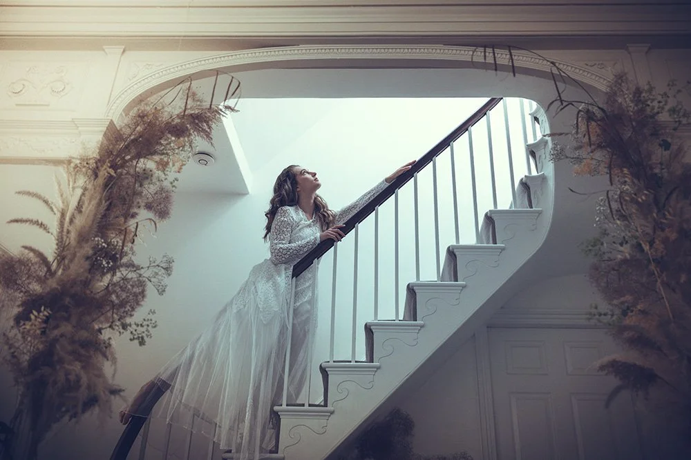Bride in white lace Tuesday's Bride wedding dress standing on staircase, looking up with hand on railing, decorated with dried plants on sides.