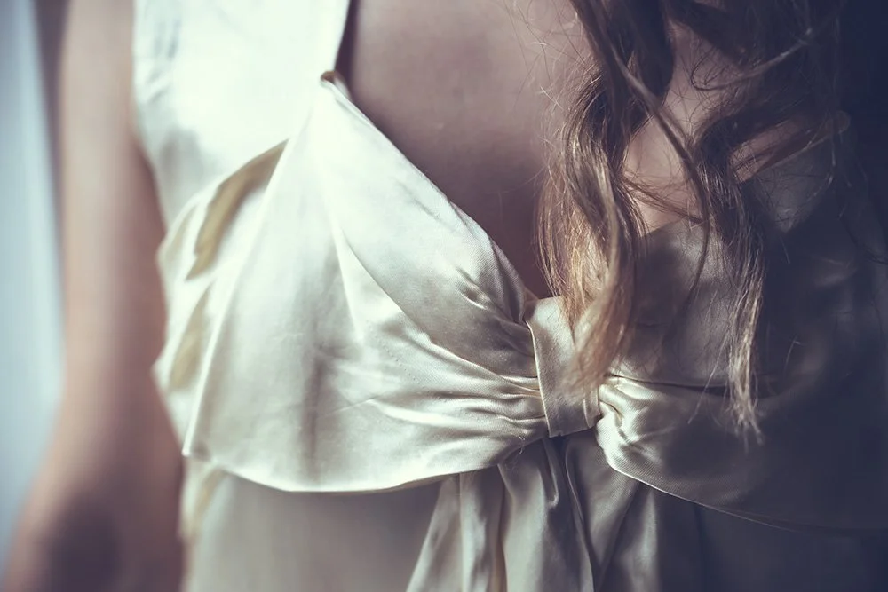 Close-up of a satin or silk light-coloured wedding dress with a large bow detail to the front, part of the bride's upper body and flowing hair visible.