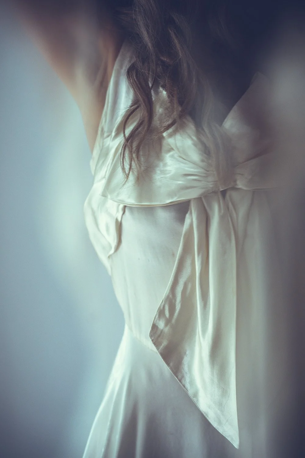 Close-up of a bride wearing a silky, satin-like white Tuesday's Bride wedding dress with a bow on the front, dark wavy hair, in front of a plain background.