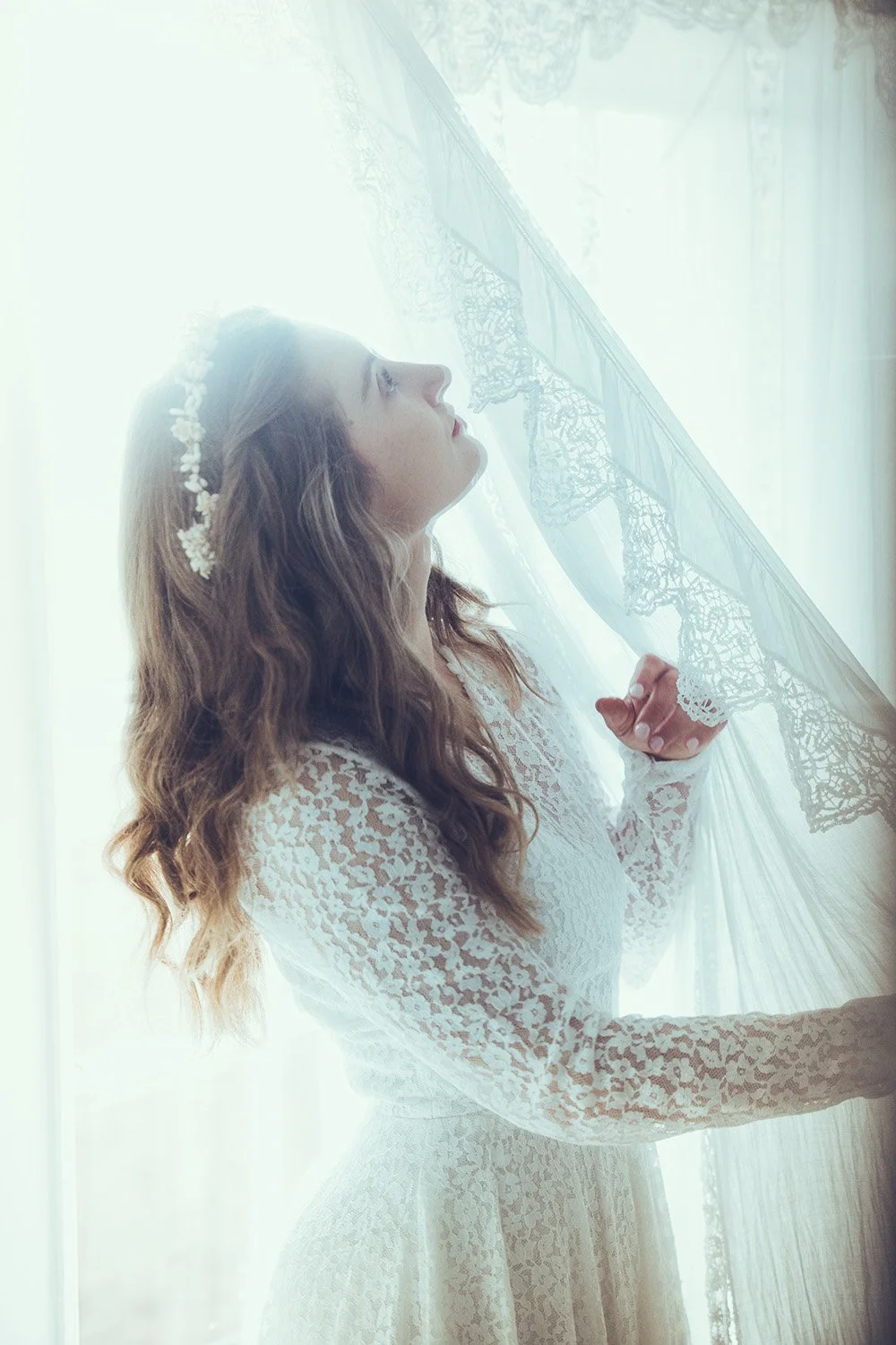 A bride in a lace wedding dress with a floral headband looks at a sheer curtain with lace trim, backlit by sunlight.