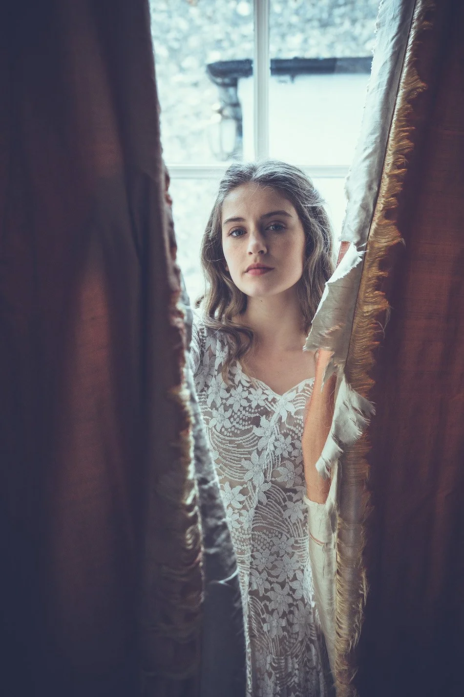 A bride with wavy brown hair and blue eyes looking through partially opened gold and brown curtains, with a window in the background.